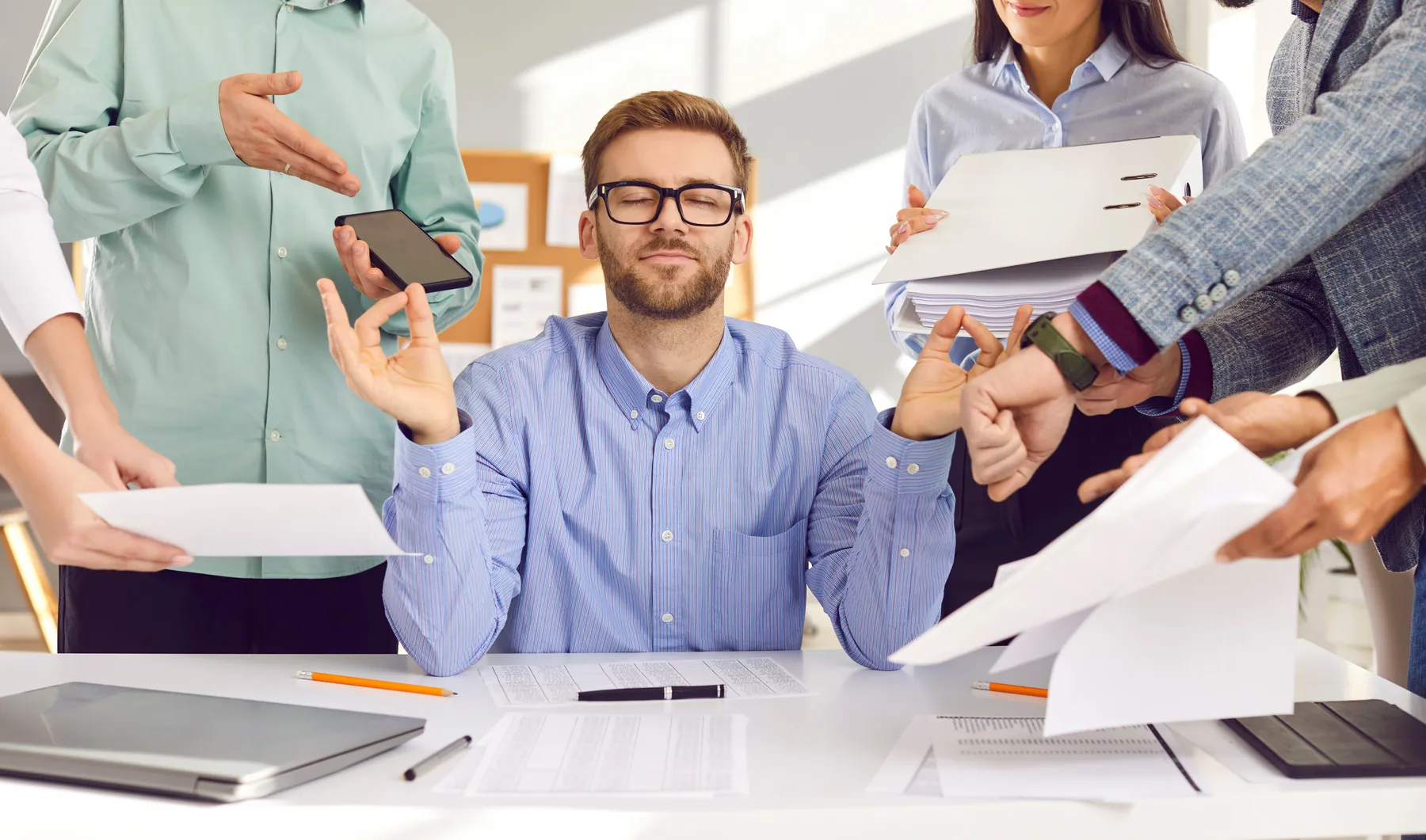 A man sitting calmly at a desk while colleagues surround him with paperwork and devices.